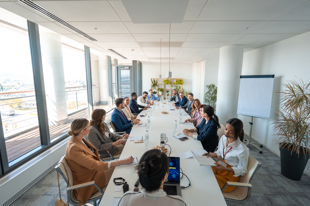 A group of diverse business executives, including men and women, attentively participating in a meeting in a modern, brightly lit boardroom with large windows and indoor plants.